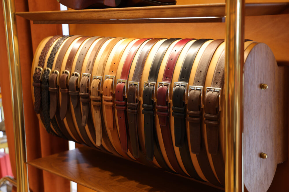 A row of leather belts in various shades of brown, black, and burgundy displayed on a wooden rack.