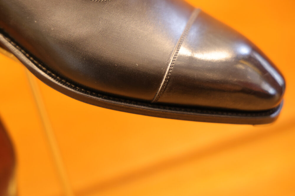 A close-up of a polished black leather shoe with a pointed toe and subtle stitching detail.