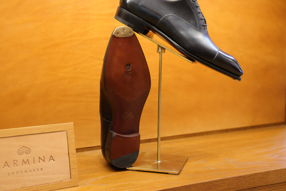A black leather shoe is displayed on a stand, showcasing its polished upper and brown leather sole.