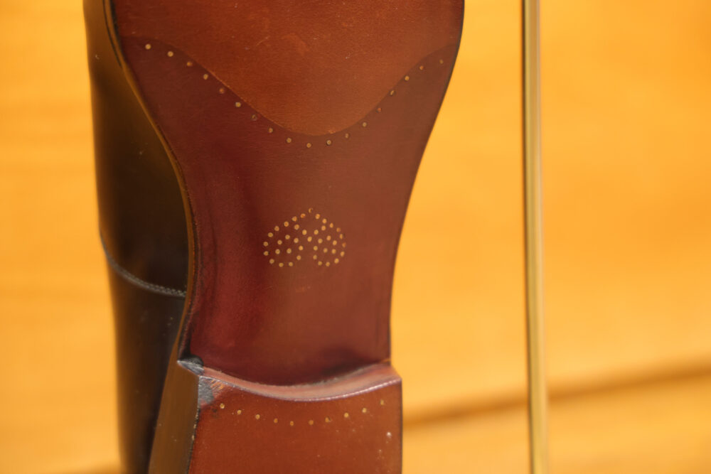 A close-up of a polished brown leather shoe heel featuring decorative perforations.