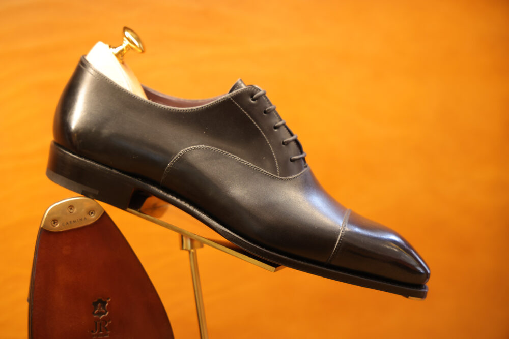 A polished black leather dress shoe is displayed on a stand with a gold shoehorn.