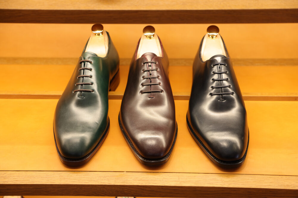 Three pairs of leather dress shoes in dark green, burgundy, and black are displayed on a shelf.