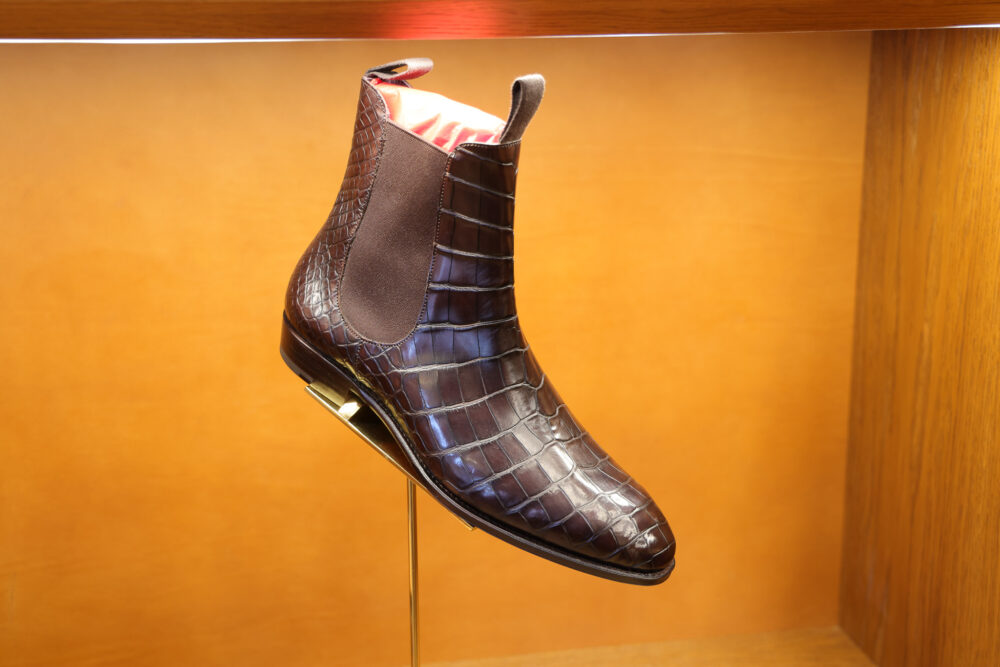 A brown crocodile leather ankle boot with elastic side panels displayed on a stand.