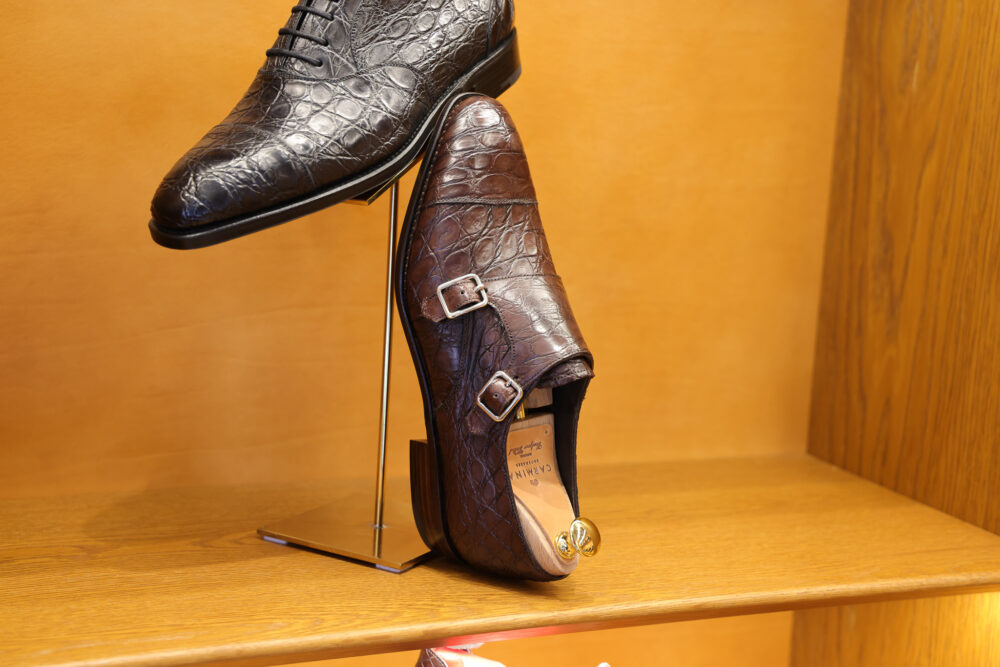 A black crocodile-patterned dress shoe and a brown double-monk strap shoe are displayed on a stand.