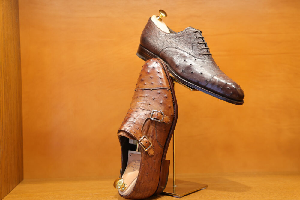 Two pairs of elegant leather shoes are displayed: a dark brown oxford shoe with perforations on top and a light brown double monk strap shoe with buckles.