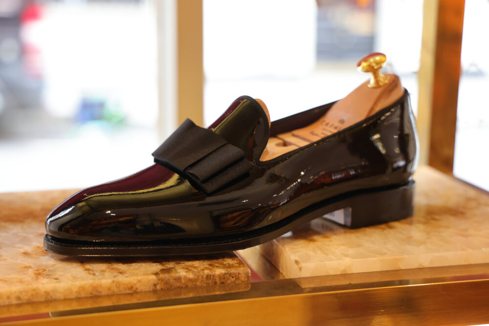 A shiny black dress shoe with a bow on the front sits on a display surface.