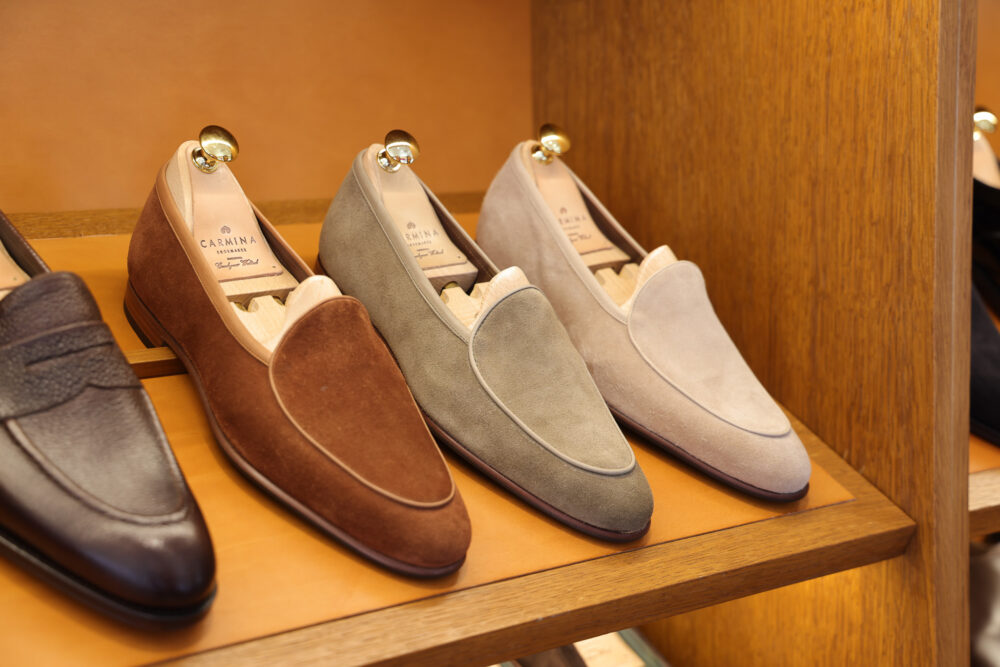 A display of three pairs of suede loafers in brown, gray, and beige, featuring gold shoehorns.