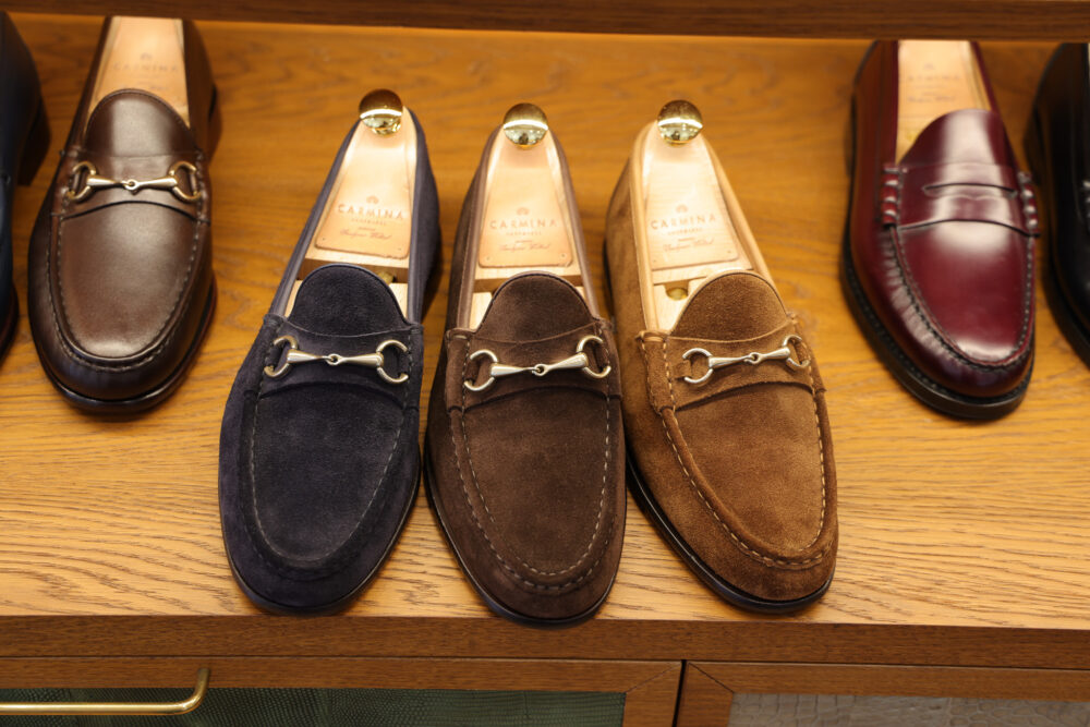 A selection of four pairs of loafers in dark brown leather, navy suede, light brown suede, and burgundy leather, featuring decorative metal accents.