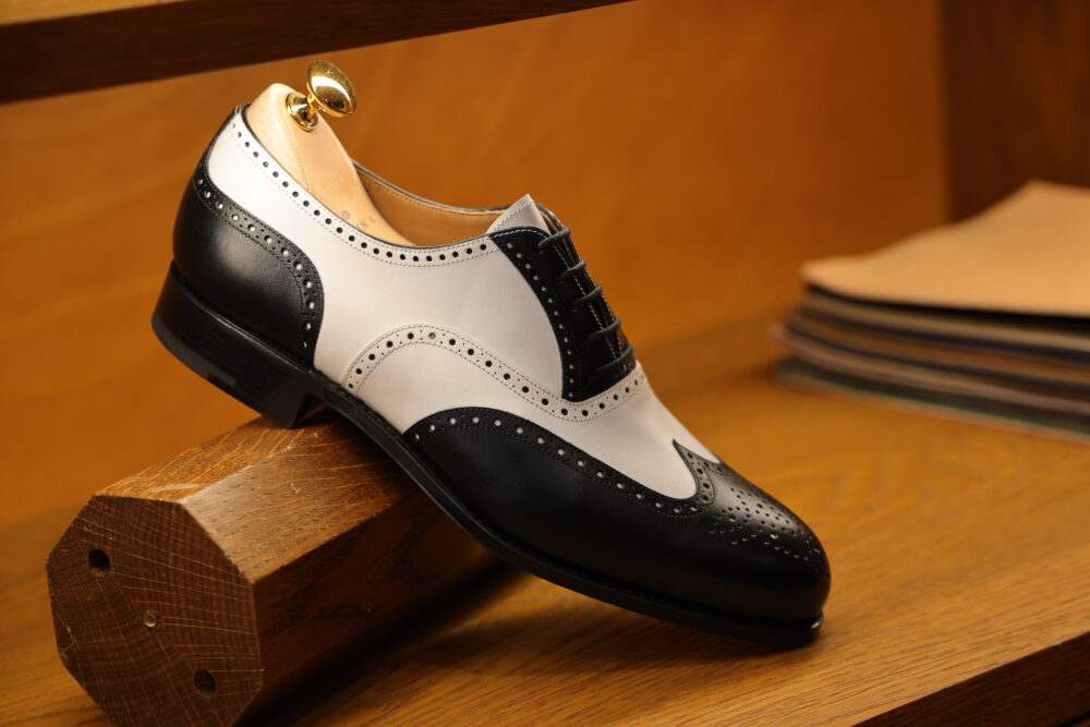 A black and white wingtip dress shoe is displayed on a wooden block.