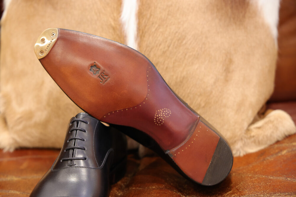 A black leather shoe with a brown leather sole featuring decorative perforations and a gold metal piece.