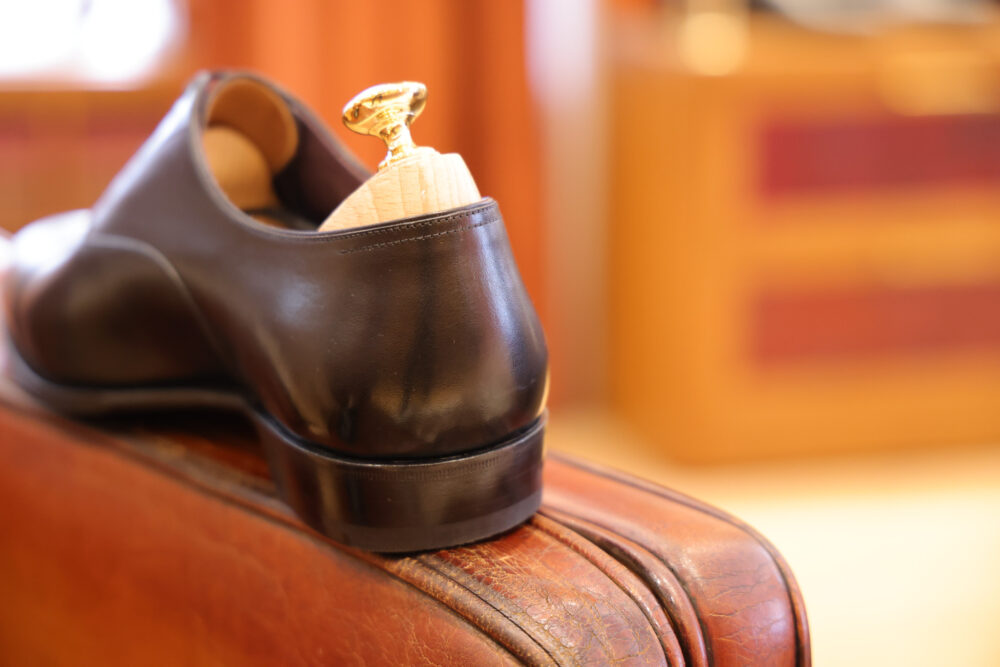 A polished black shoe with a wooden shoe tree and gold knob resting on a brown leather suitcase.