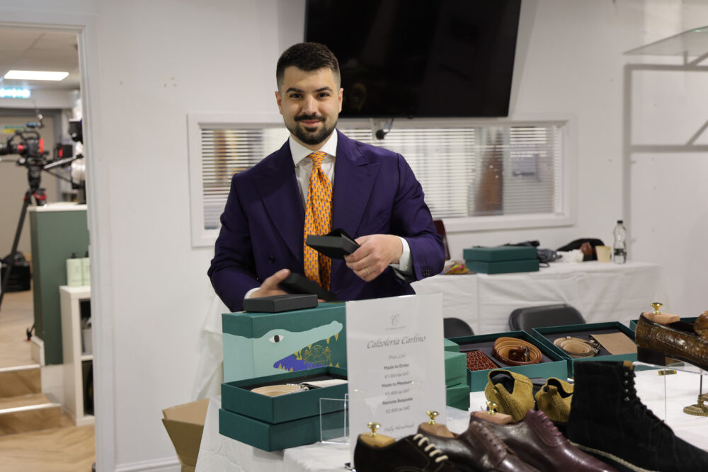 A man in a purple suit and orange tie is holding a black box at a table displaying various shoe boxes and accessories.
