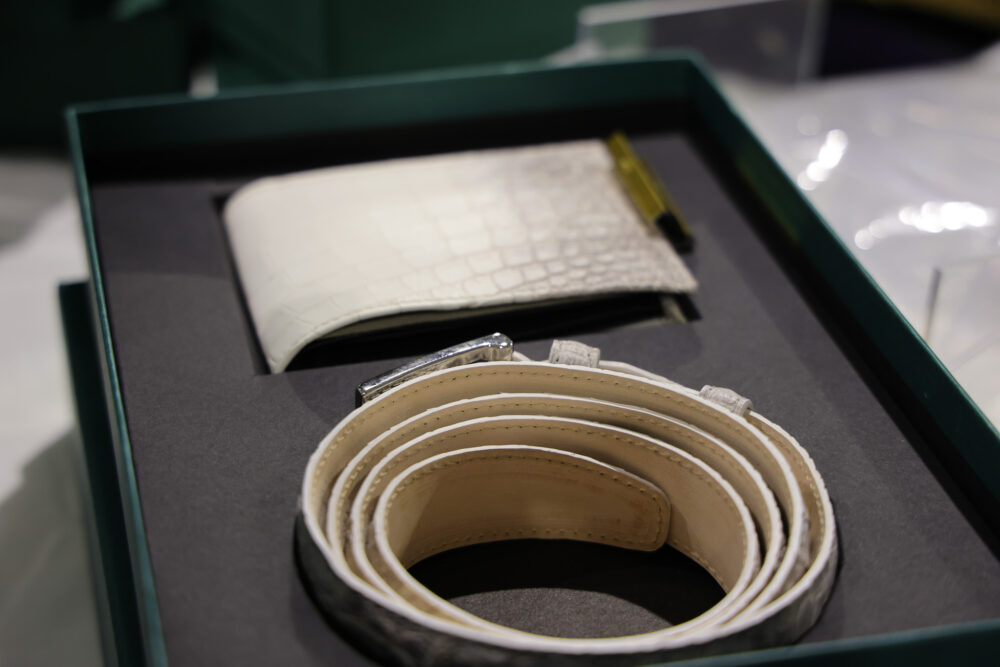 A white crocodile-textured wallet and a rolled light-colored leather belt are displayed in a dark box.