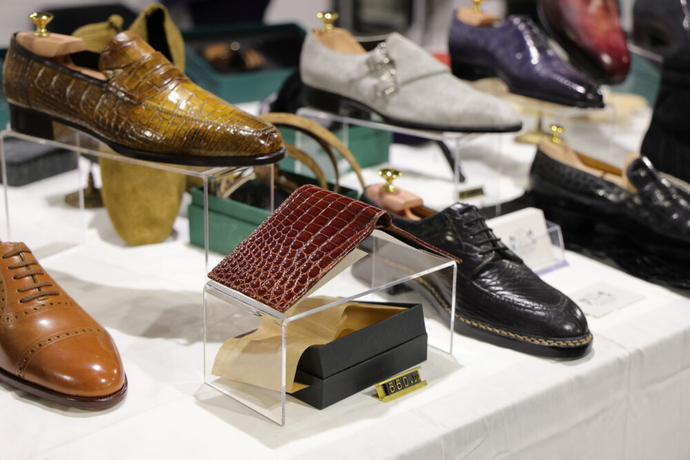 A display of various leather shoes and a burgundy crocodile-patterned wallet in a clear case.