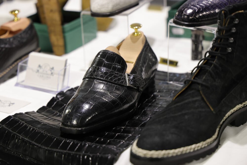 A polished black crocodile leather shoe with a strap and buckle is displayed on a textured black leather surface.