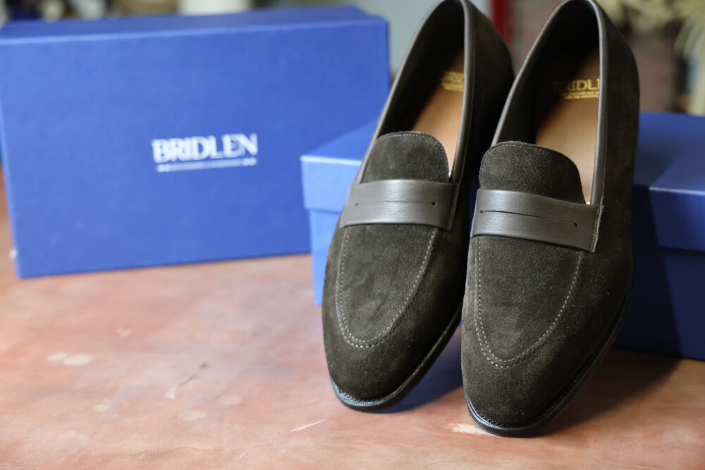 A pair of dark green suede loafers with brown leather accents, placed in front of a blue box.