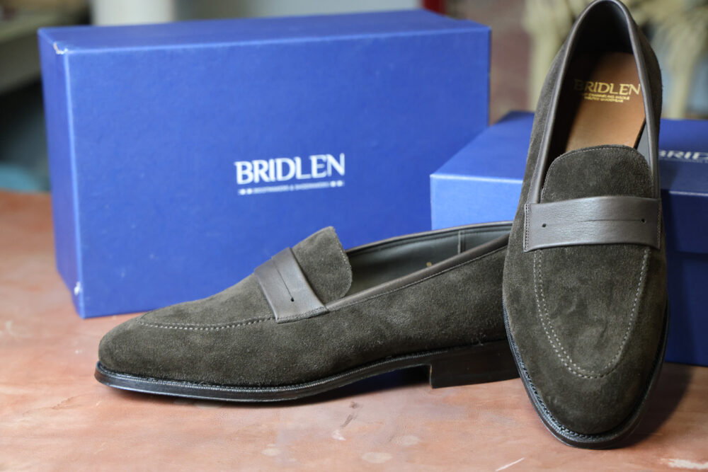 A pair of dark green suede loafers with a brown leather strap, next to a blue box labeled "Bridlen."