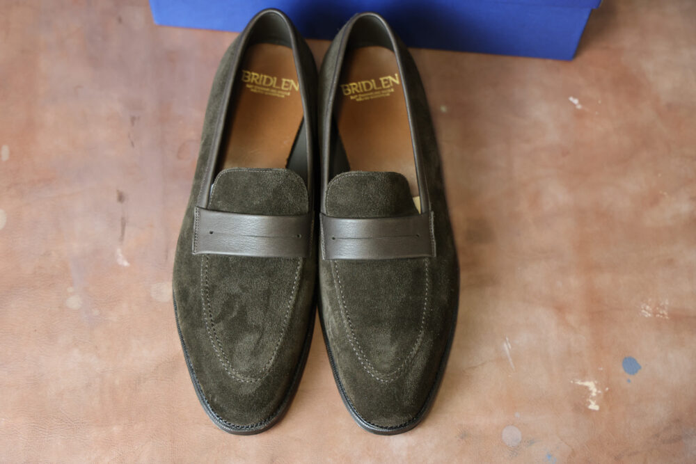 A pair of dark green suede loafers with a brown leather strap across the top.