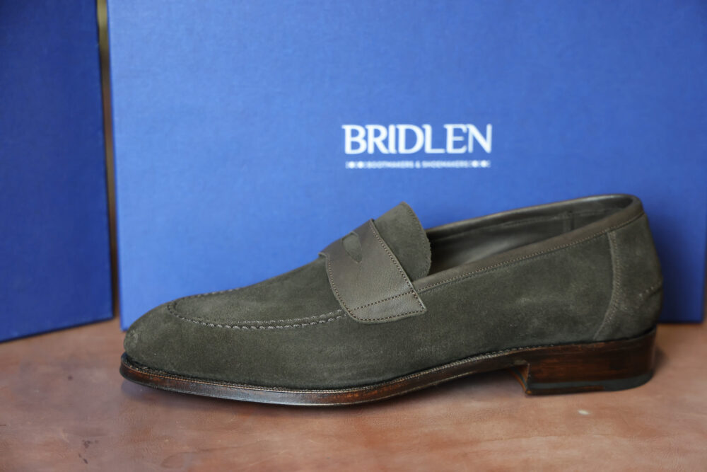 A dark green suede loafer shoe with a leather strap across the front.