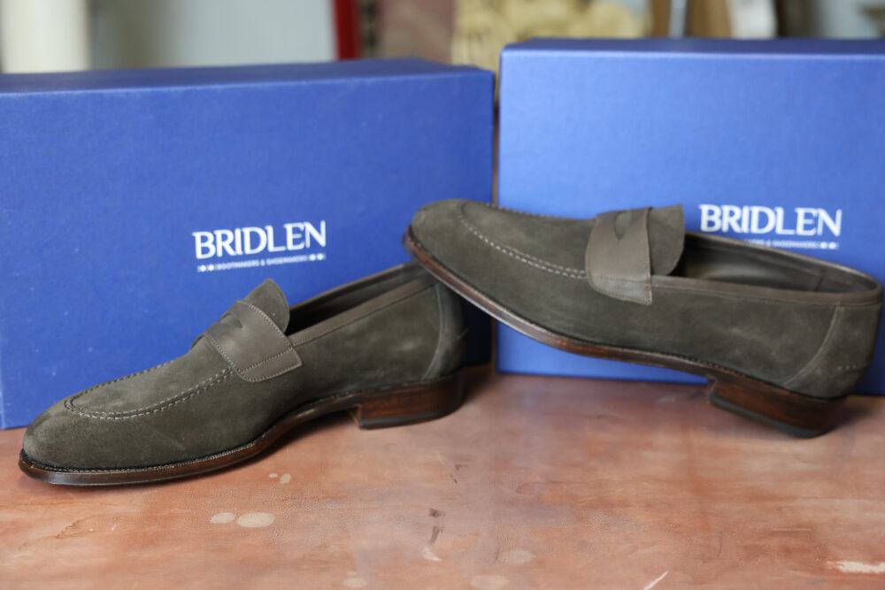 A pair of dark green suede loafers with leather accents, placed near blue boxes labeled "BRIDLEN."