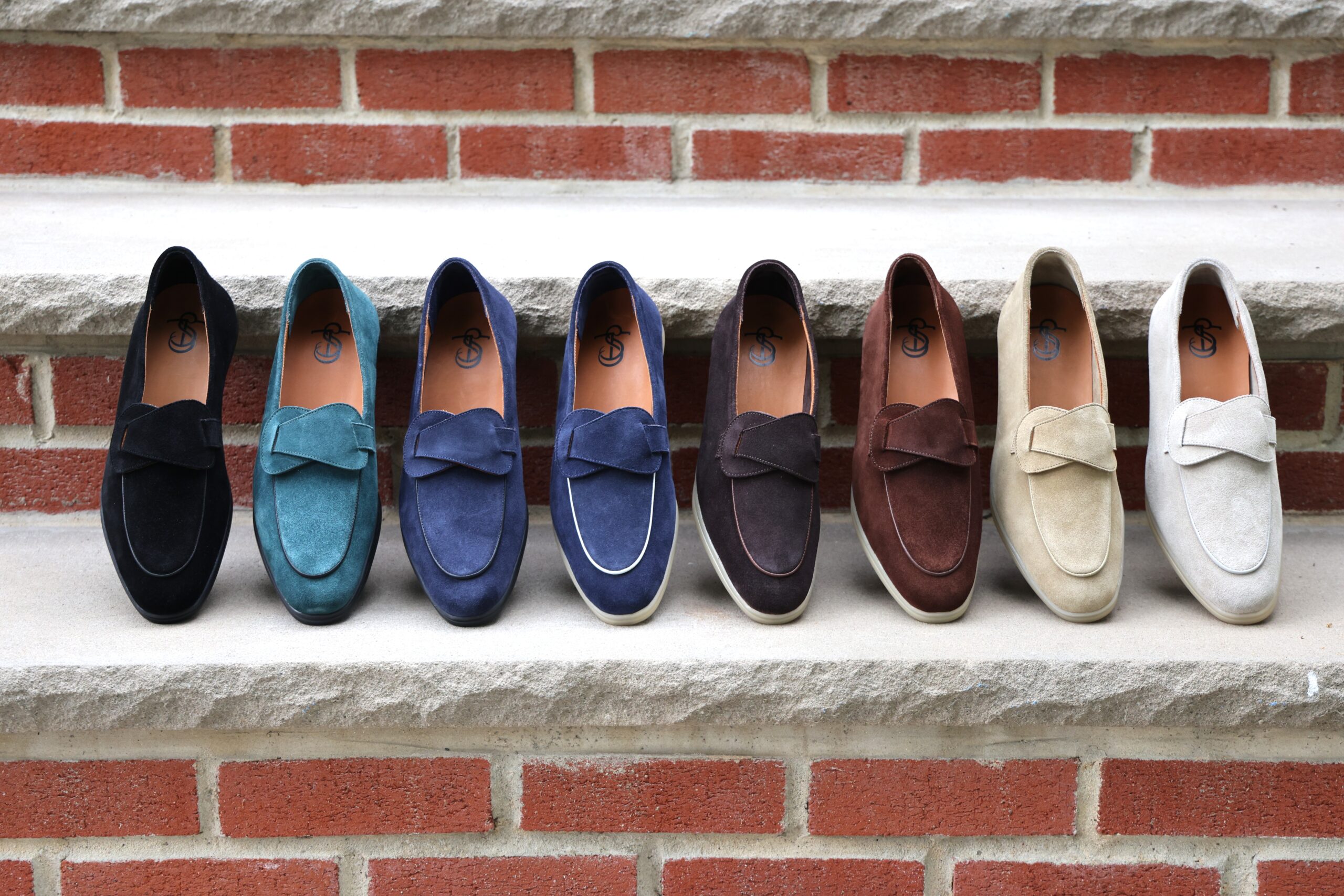 A row of eight pairs of suede loafers in black, teal, navy, brown, and cream, featuring a bow detail on each pair.