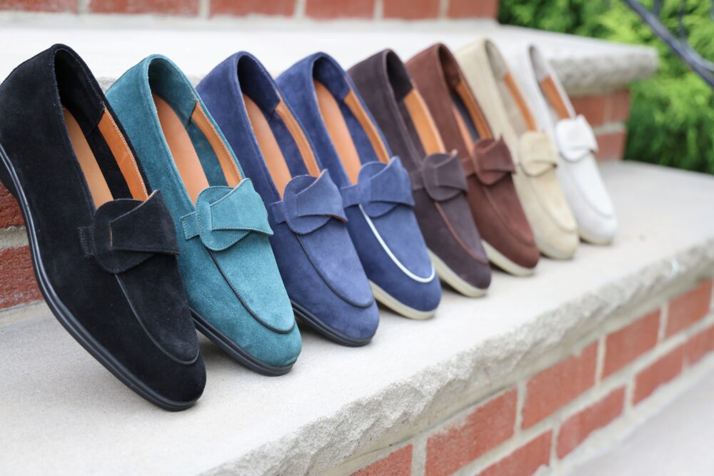 A row of eight pairs of suede loafers in black, teal, navy, brown, and beige, each featuring a bow detail.