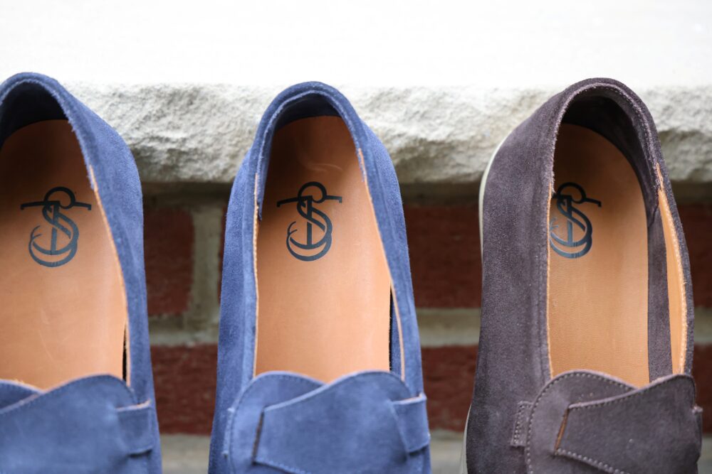 Three pairs of suede loafers in navy blue and dark brown, with leather insoles featuring a logo.