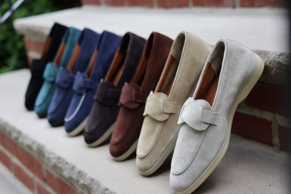 A row of suede loafers in various colors, including black, blue, green, brown, beige, and gray, displayed on a ledge.