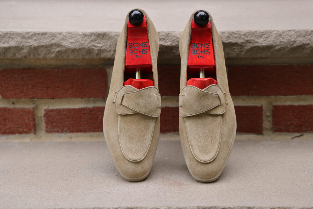 A pair of beige suede loafers with red insoles and wooden shoe trees.