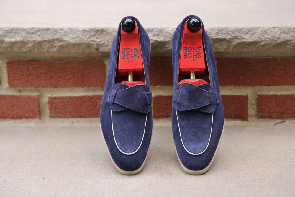 A pair of navy blue suede loafers with white piping and red shoe trees inside.