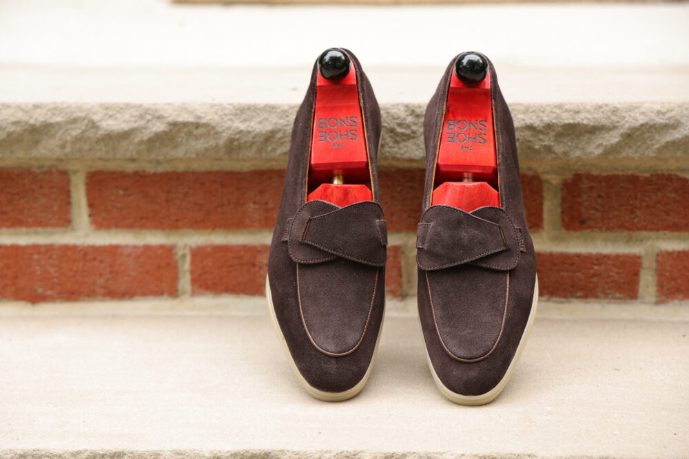 A pair of dark brown suede loafers with red insoles and wooden shoe stretchers.