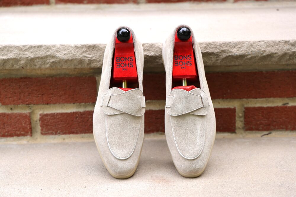 A pair of light beige suede loafers with a decorative strap and red insoles.