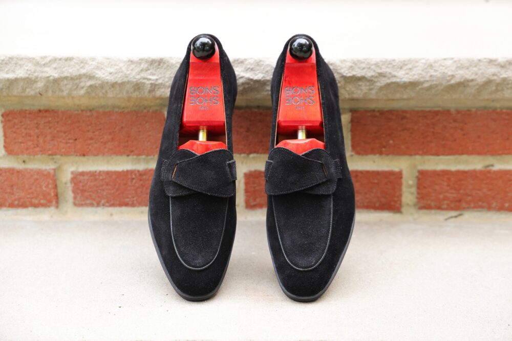 A pair of black suede loafers with a decorative bow and wooden shoe trees inside.
