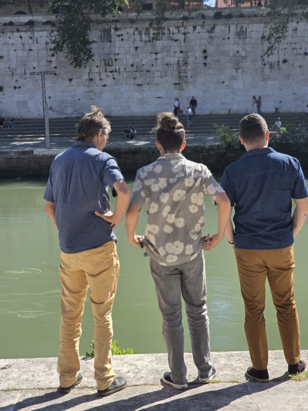Three people, wearing blue, patterned, and dark shirts with beige, gray, and brown pants, stand by a green river.
