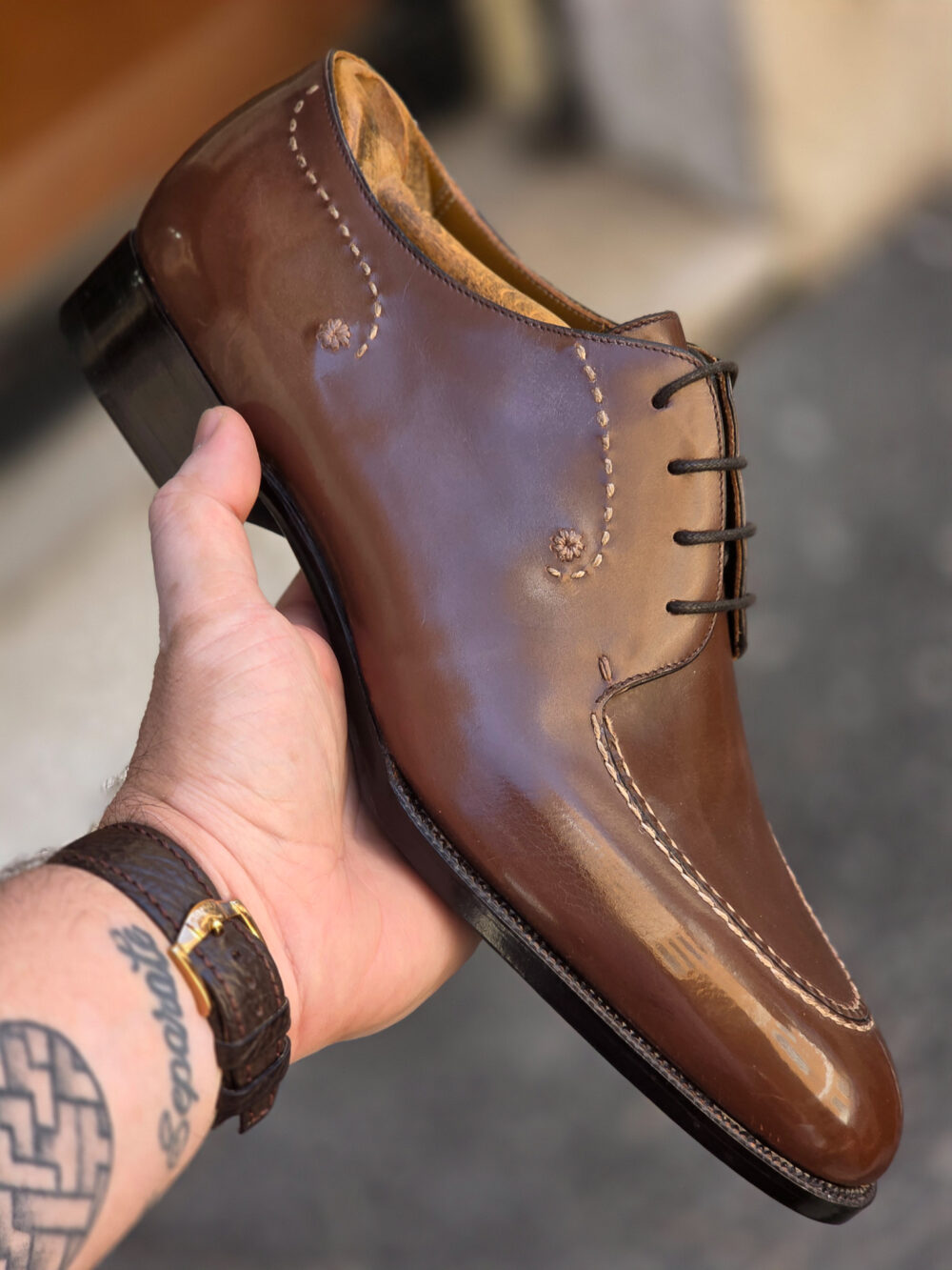 A hand holds a polished brown leather shoe with decorative stitching and a black sole.