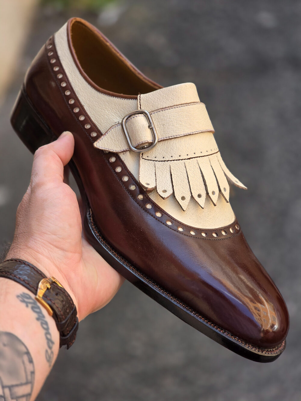 A brown and cream leather shoe with decorative fringe and a buckle, held in a hand.