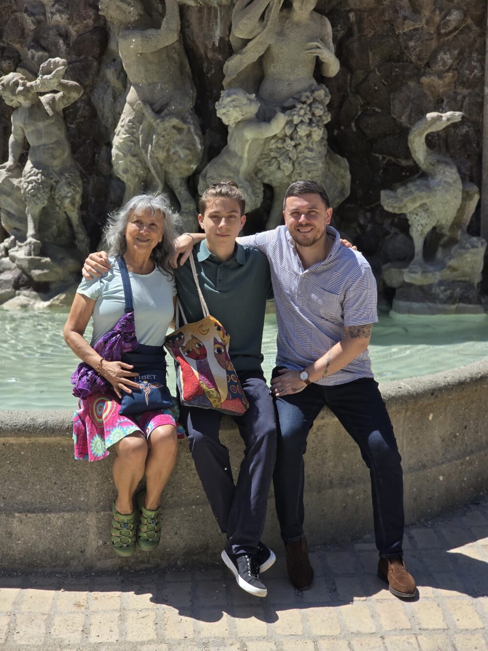 Three people sit together, a woman with gray hair in a colorful skirt, a young man in a green shirt, and a man in a striped shirt, all smiling.