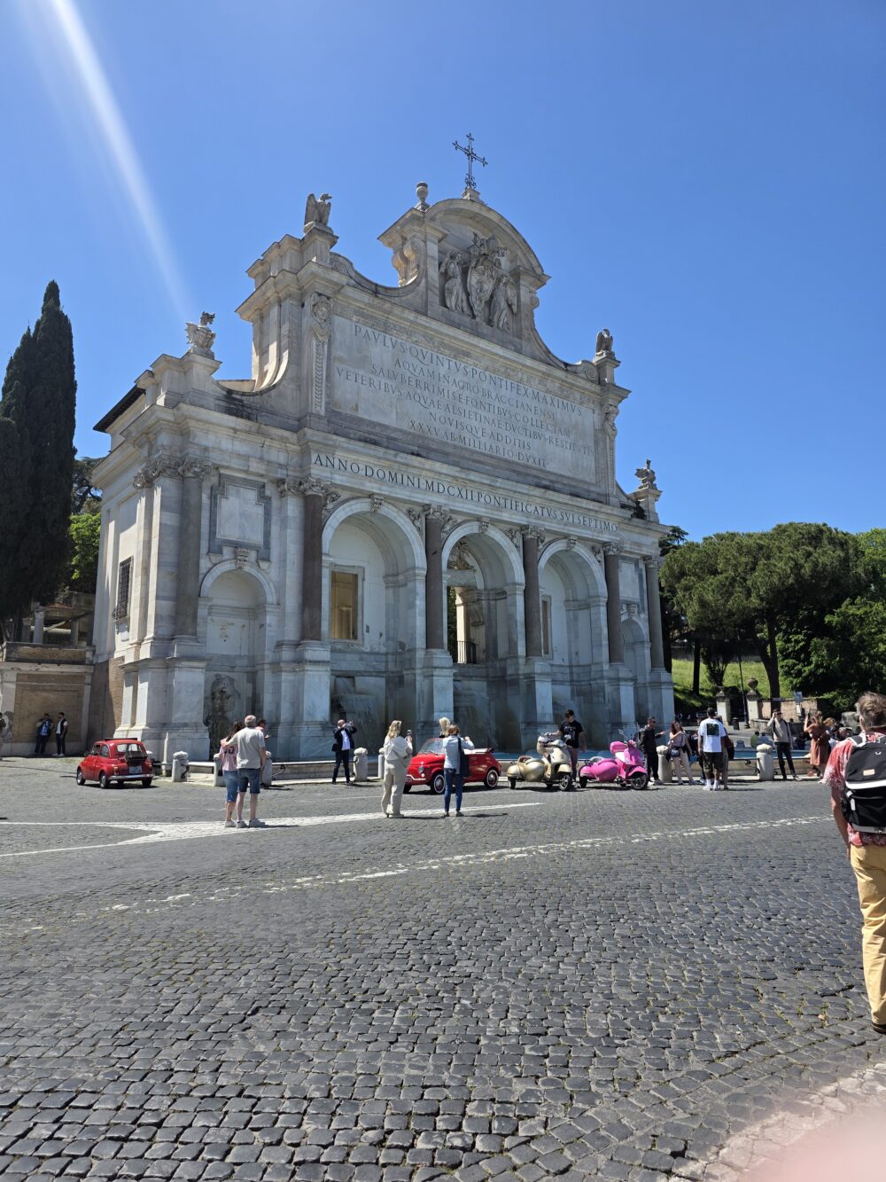 A large stone building with a cross and sculptures at the top, featuring inscriptions and surrounded by people and small colorful cars.