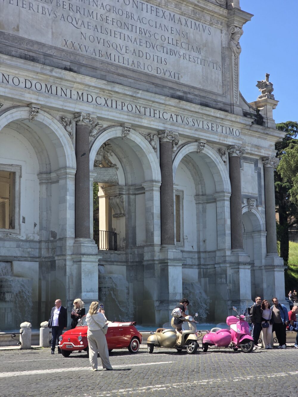 A woman takes a photo in front of a historic arch, with a red car and gold and pink scooters nearby.