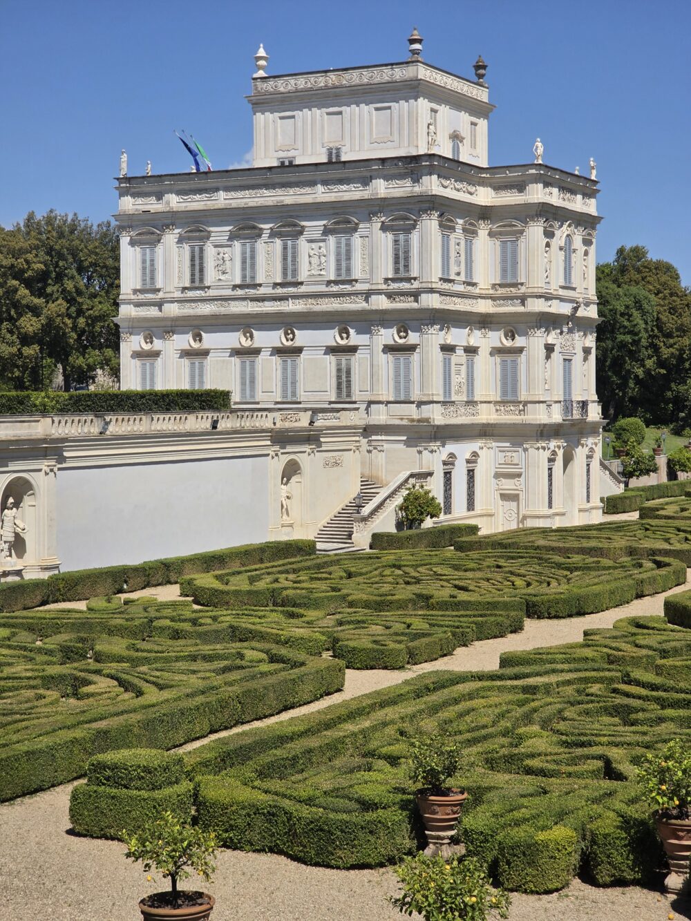 A white ornate building with decorative details stands beside a neatly trimmed hedge maze.