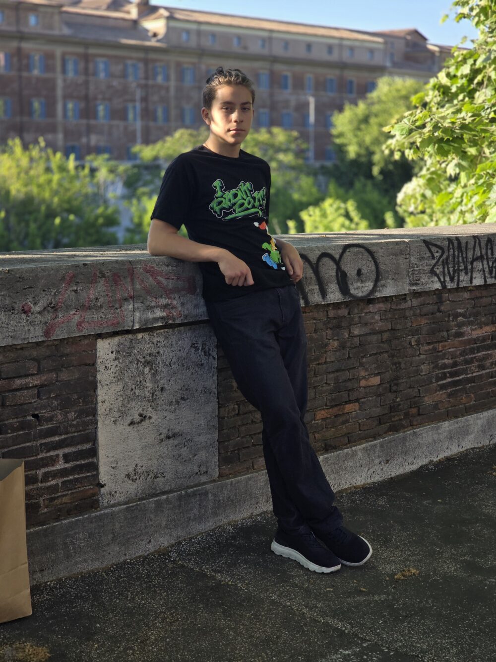 A young person with long hair, wearing a black t-shirt featuring green text and a cartoon character, leans against a stone wall.
