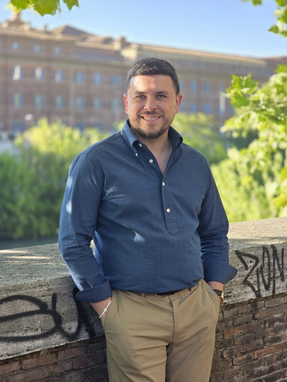 A man with short dark hair smiles while wearing a blue button-up shirt and beige pants, standing by a stone wall.