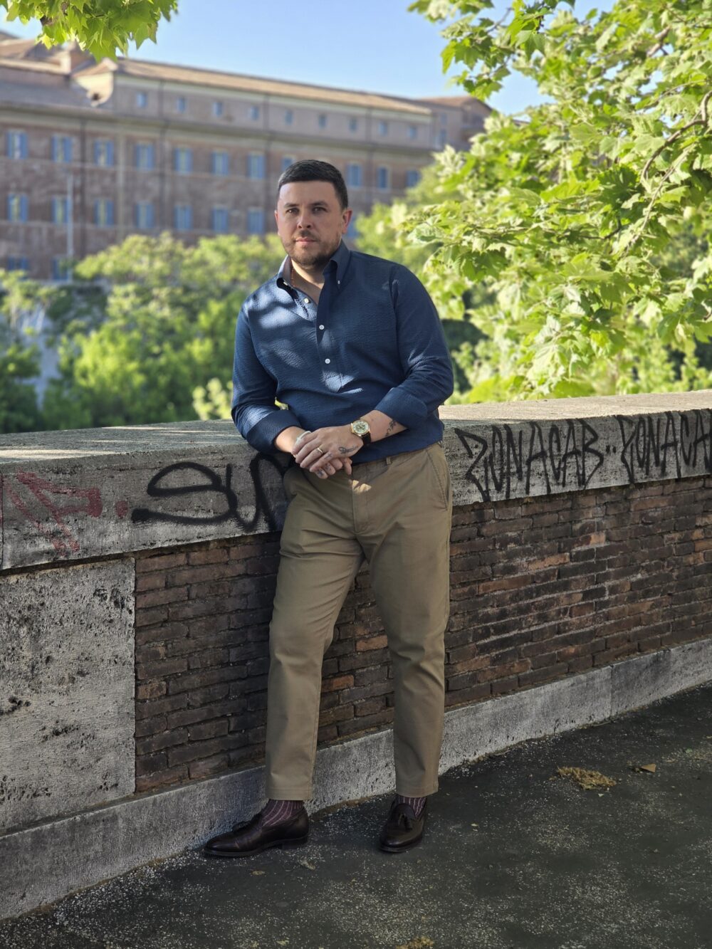 A man in a dark blue shirt and khaki pants leans against a brick wall.