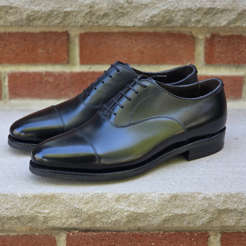 A pair of polished black leather dress shoes with a cap toe design and laces.