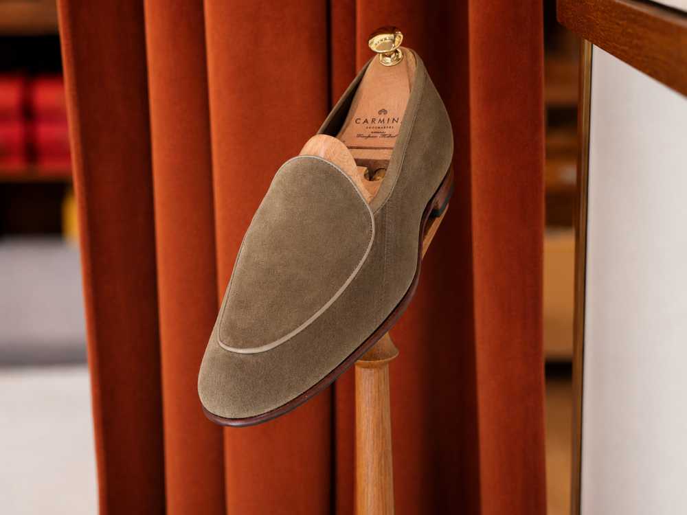 A green suede loafer shoe displayed on a wooden stand.