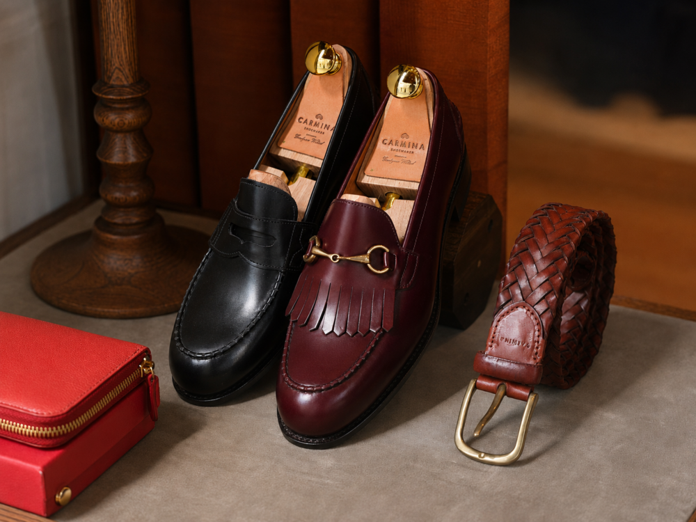 A pair of leather loafers, one black and one burgundy with a gold accent, alongside a braided brown belt and a small red wallet.