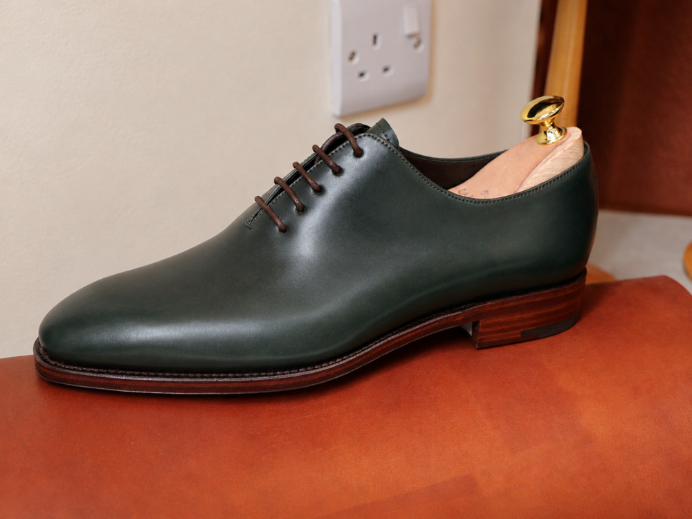 A dark green leather dress shoe with brown laces and a wooden heel.