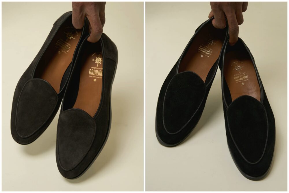 A hand holds a pair of black suede loafers with brown insoles.