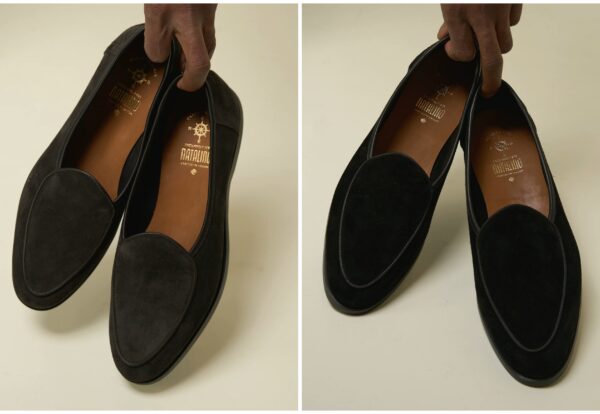 A hand holds a pair of black suede loafers with brown insoles.