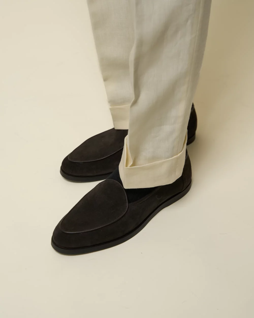 A pair of dark brown suede loafers worn with light-colored trousers.